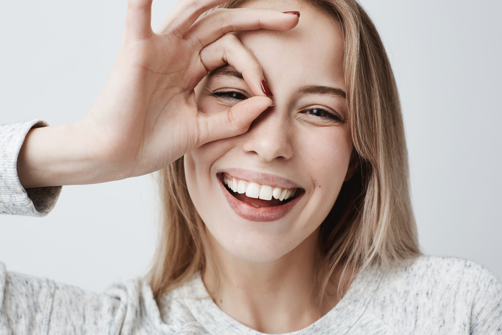 Dental Implants Clairemont: Bright Smile Confidence Laughing woman making an OK hand gesture over her eye, showing the kind of bright smile dental implants in Clairemont patients hope to achieve